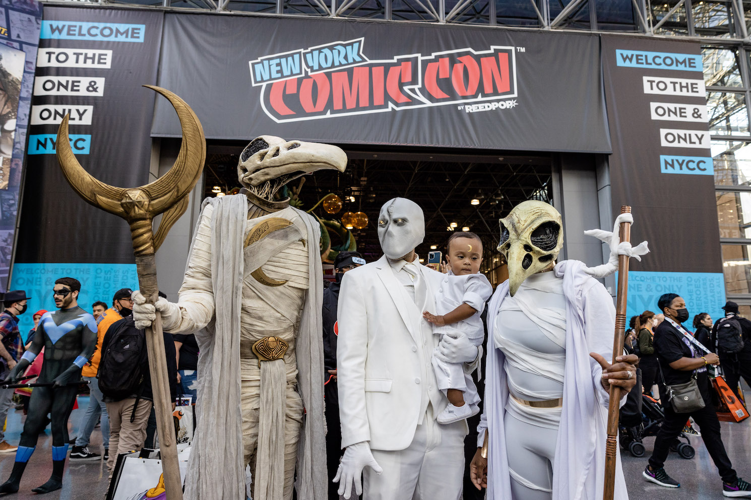 A family of four dressed in Moon Knight costumes. On the left, a person holding a staff with a crescent moon on the top of it wears mummy wrappings and a bird skull on the head. In the center, a person in a white suit and mask with a crescent moon holds a baby in a matching suit. On the right, a cosplayer in a white bodysuit, wrapped in mummy wrappings holds a brown staff.