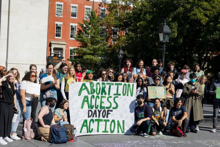 A group of protesters poses in front of the Washington Square Arch for a photo. In the middle is a banner that reads “ABORTION ACCESS DAY OF ACTION”.