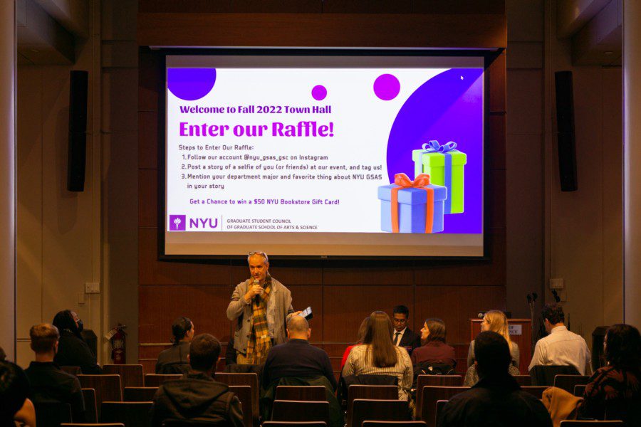 A male holding a microphone standing among a group of people sitting in a conference hall with a projector screen in the back indicating that this is the Fall 2022 Town Hall event of NYU’s Graduate School of Arts and Science.