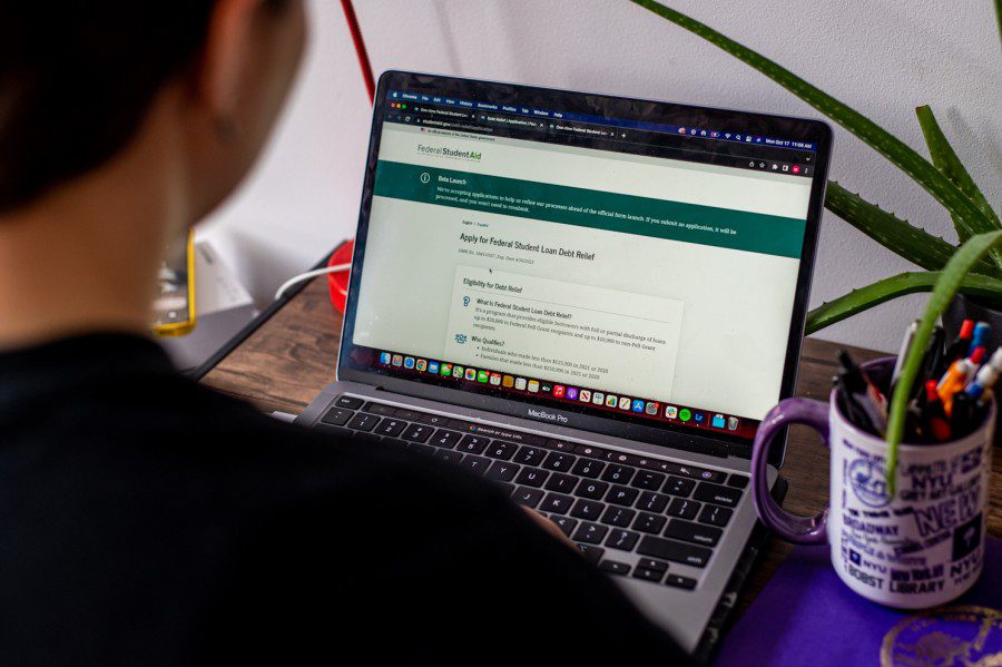 The back of a woman wearing a black t-shirt looking at a computer screen that reads “Apply for Federal Student Loan Debt Relief.”