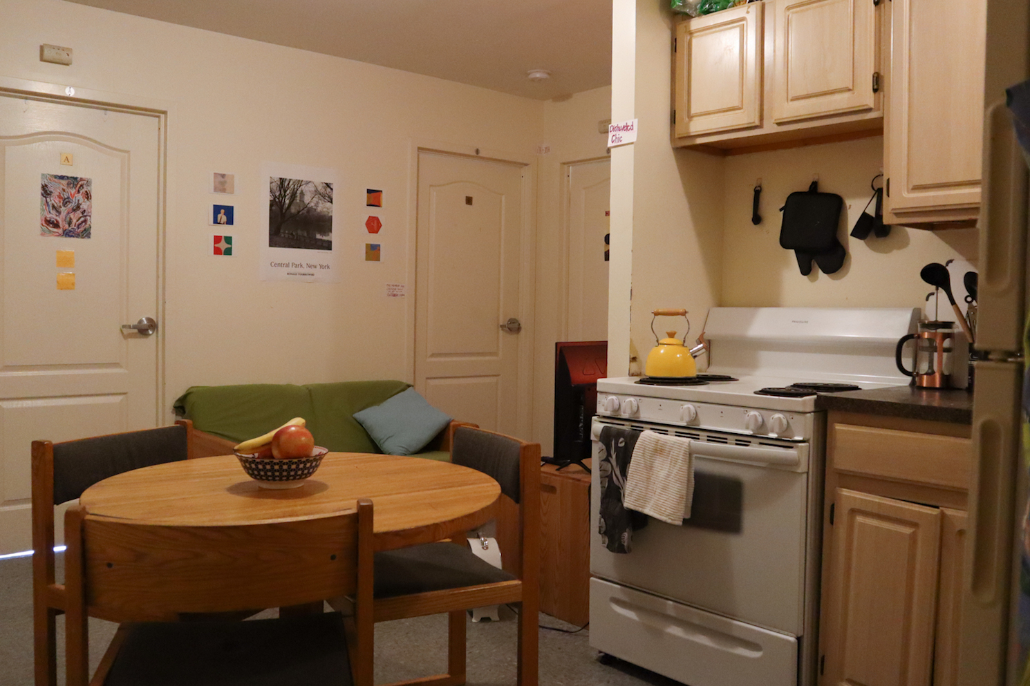 A dorm kitchen with a wooden dining table and stove top. A fruit basket is on the table and a yellow kettle is on the stove.