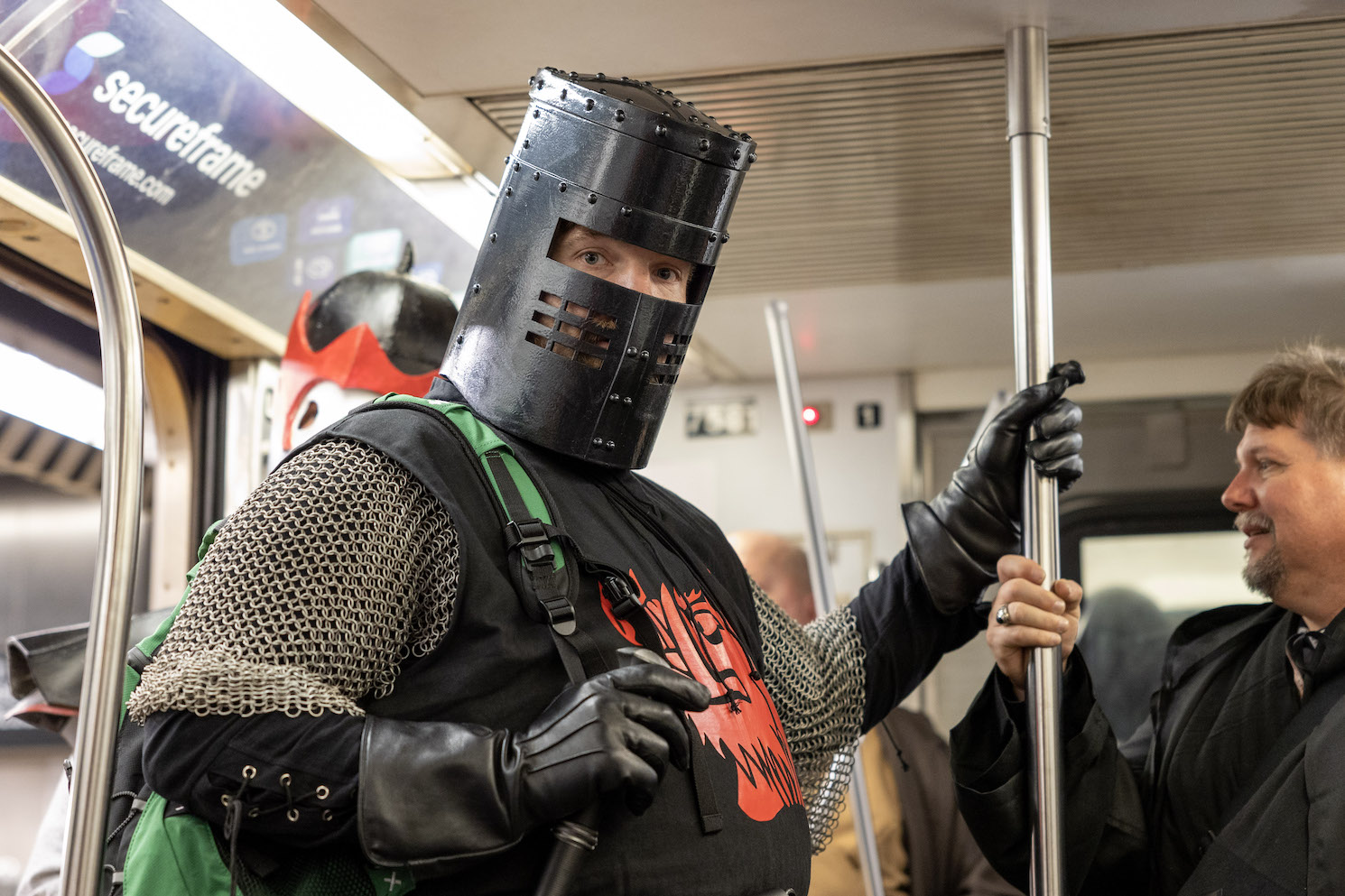 A subway passenger dressed in a black-and-silver knight costume with a red dragon-like figure on his stomach wears a black cylinder over his head. The head covering has a rectangular hole for his eyes, and small rectangular holes around his nose and mouth.