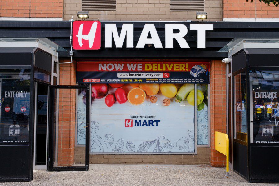 The storefront of the H Mart on 39 third Avenue in Manhattan with a poster of fruits and vegetables and text “WE NOW DELIVER!” under the H Mart logo.