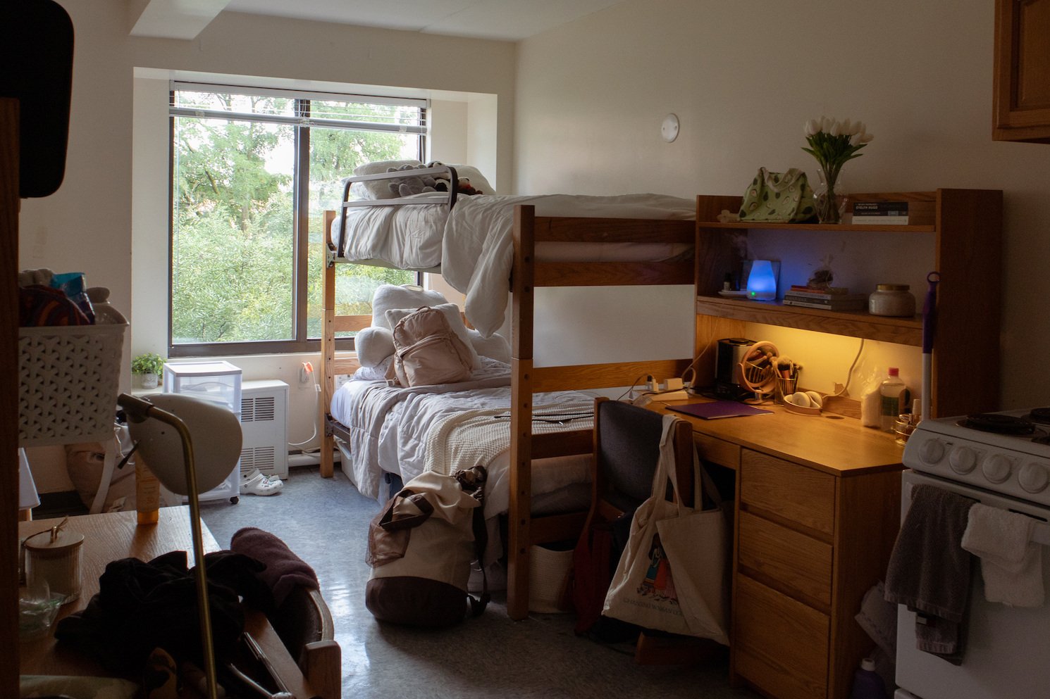 A dorm room bunk bed with white sheets next to a wooden desk. In the background, a window.