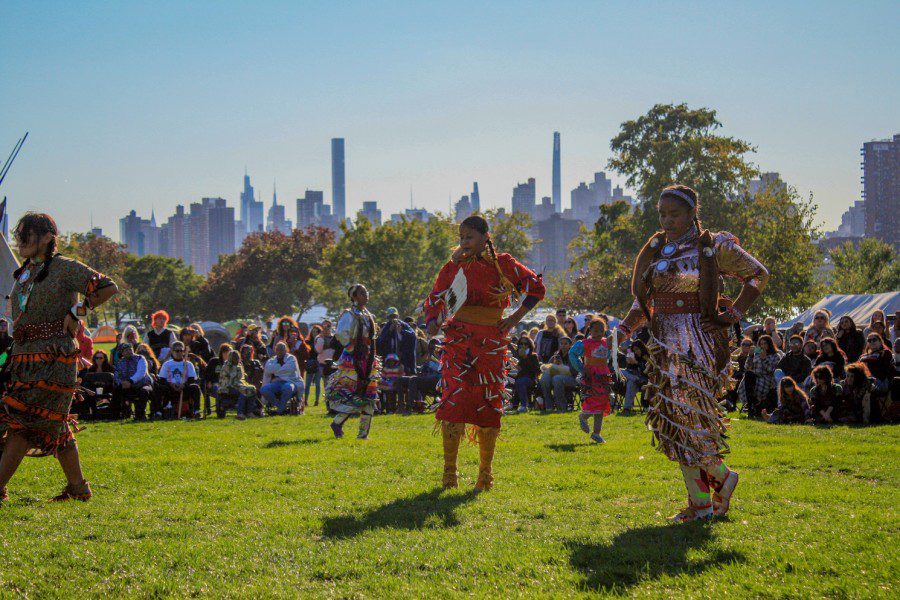 Three women wearing jingle dresses dance around a smoking fire pit on Randall’s Island.