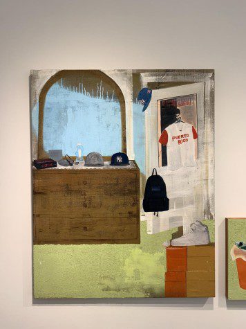 A bedroom with a green floor. To the right, a wood dresser with items on top of it, including a Yankees baseball cap. Next to the dresser, hanging from the door of the room, a red-and-white baseball jersey that reads “Puerto Rico.”