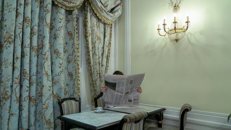 A person sits at a table, holding a newspaper in a blue-and-gold adorned room.