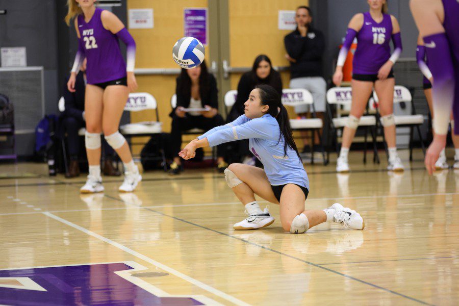 Two members of NYU’s women volleyball team pass a volleyball ball through the net to the opposing team dressed in red.