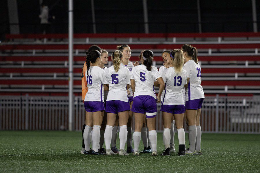 NYUs women’s soccer team wearing white jerseys, purple shorts and white socks huddles on the field.