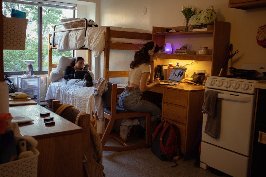 A dorm room with a girl laying down in the lower bunk bed. Another girl sits at an adjacent desk, wearing blue jeans, a white t-shirt, and pink headphones. The girl at the desk is looking at her computer screen.