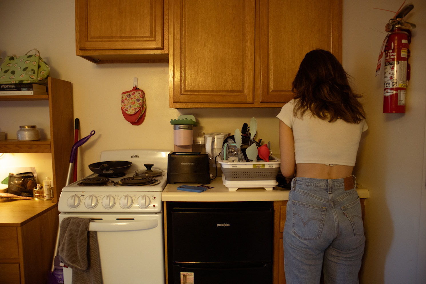 The back of a girl wearing blue jeans and a white t-shirt. She stands in a kitchen with a white oven and three wooden cabinets.