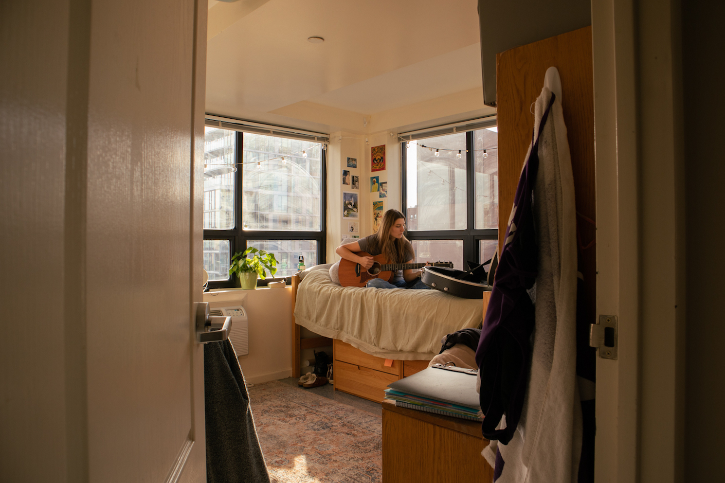 A girl sitting on a dorm bed playing a guitar.