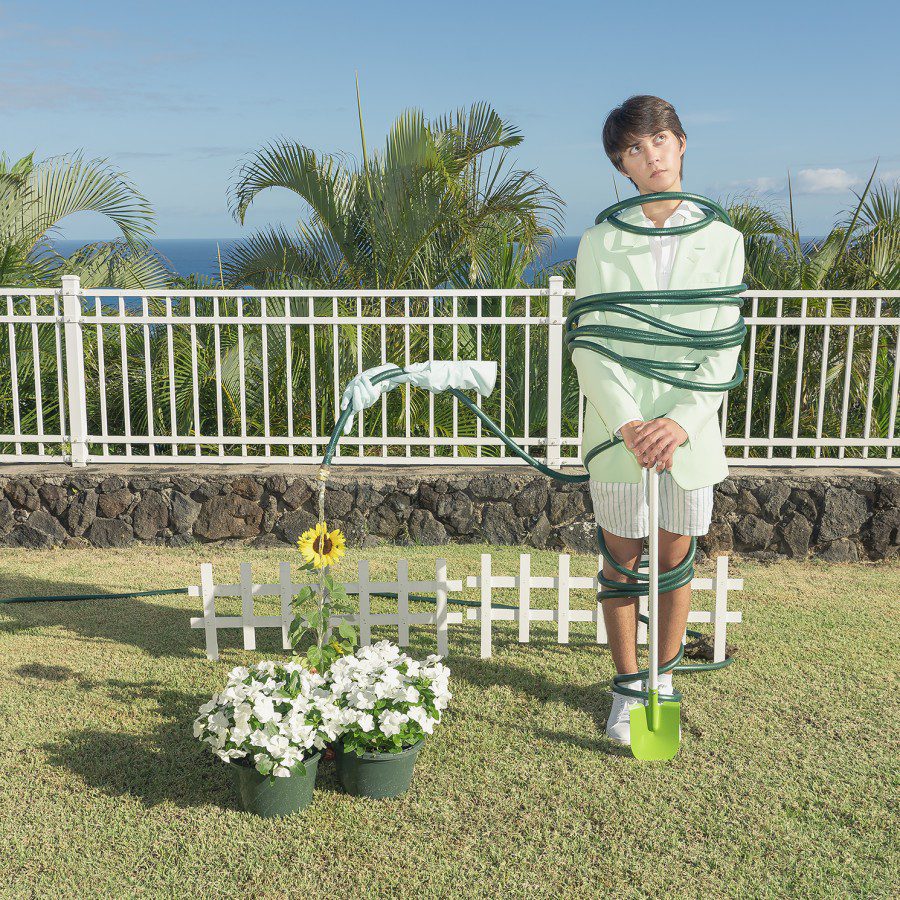Photograph of a boy holding a bright green shovel standing in a garden overlooking the ocean with a floating hose wrapped around him. The boy is watering a bed of flowers. A white fence, palm trees and a blue sky are in the background.