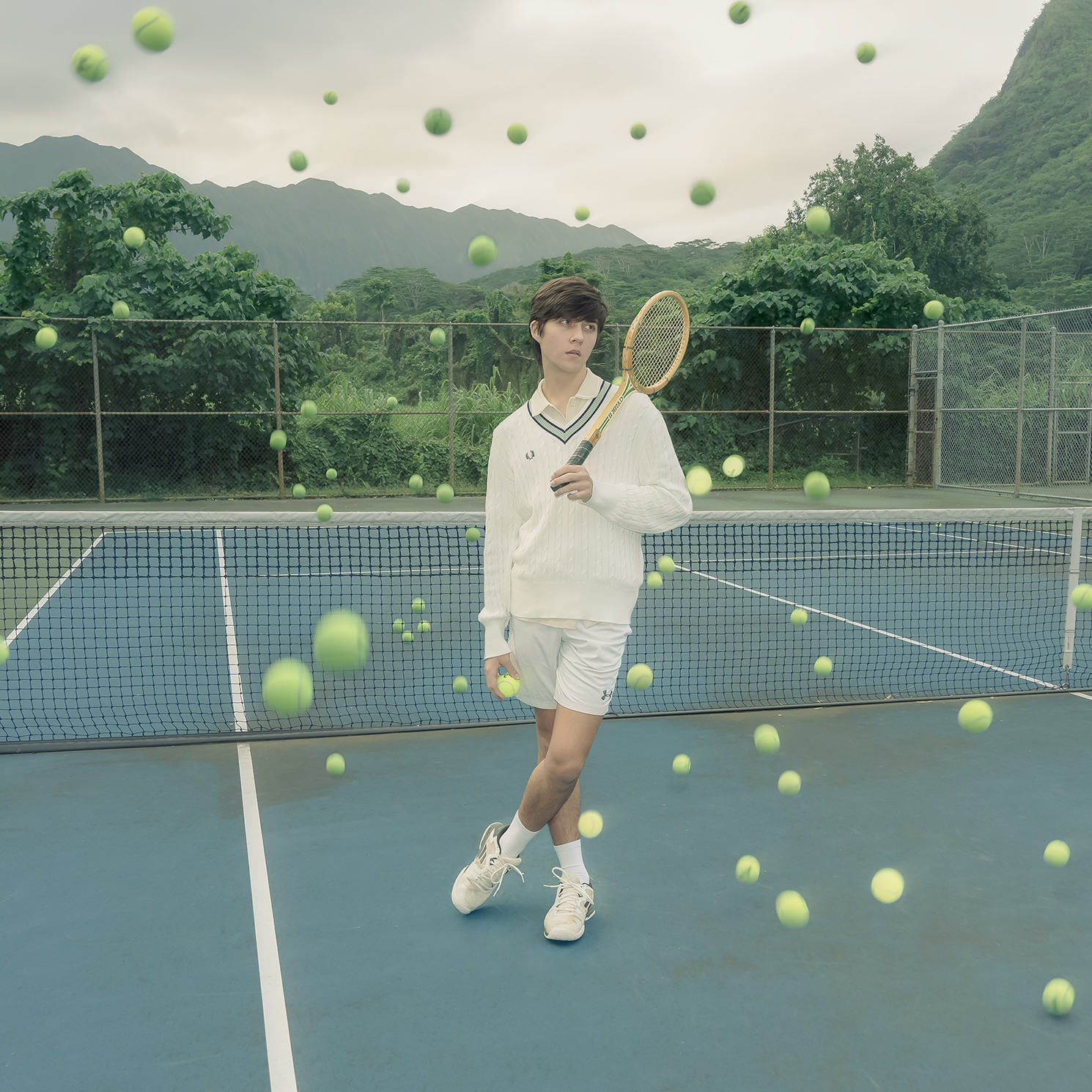 Photograph of a boy holding a tennis racket in a blue tennis court surrounded by several floating tennis balls. There are trees and mountains in the background.