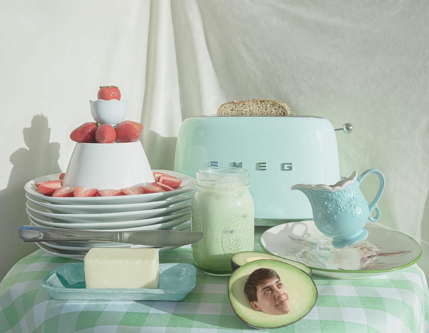 Photograph of a breakfast spread on a table with a green-and-white gingham tablecloth. On the table is a Smeg toaster with bread inside, a tower of strawberries, an iced matcha latte, a stick of butter with a floating knife, a floating tea cup, and an avocado with a boy's head as the seed. A beige fabric backdrop is behind the table.