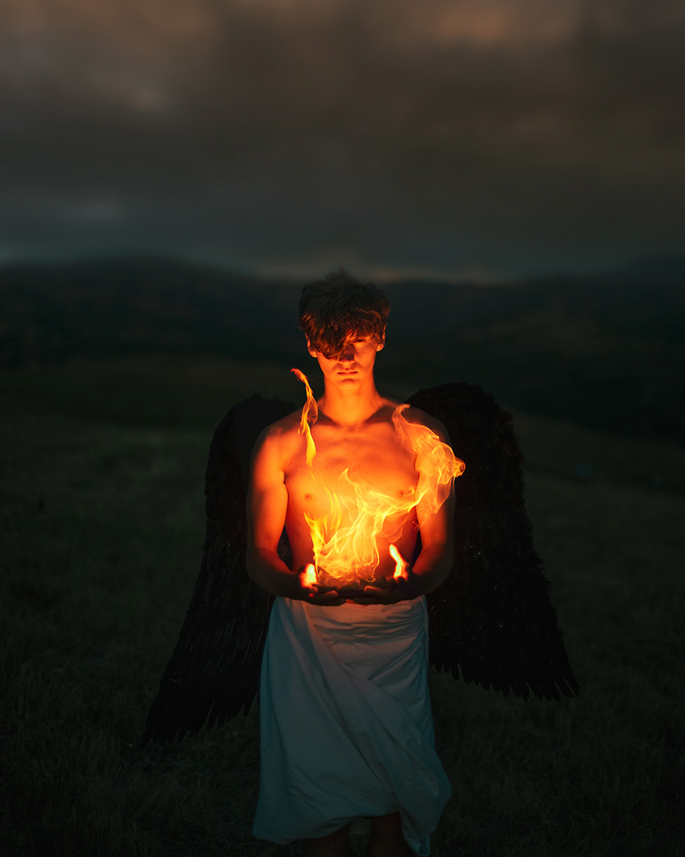 Photograph of a boy with black angel wings standing in a dark, open field holding fire in his hands. The boy is illuminated orange by the glow of the fire.