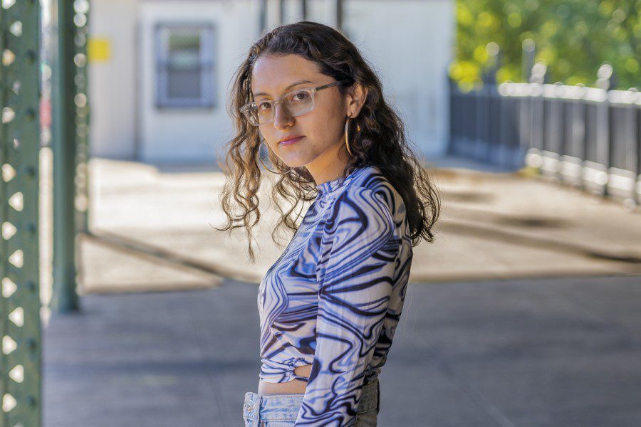 Lorraine Olaya looks at the camera while standing outside on the platform of a train station. Lorraine is wearing a patterned blue shirt and light blue jeans.