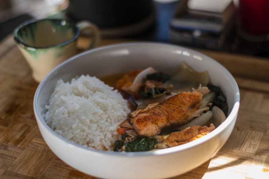 A bowl of rice and Sinigang, a Filipino soup made of fish and vegetables, sit in a bowl on a counter with a mug in the background.