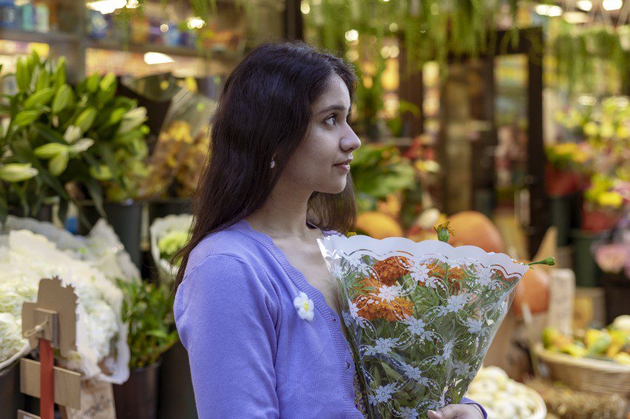 Sara Sharma holds a bouquet of orange flowers while standing in front of a deli displaying flowers in Union Square. Sara is looking into the distance, and is wearing a white floral dress and a lilac cardigan.