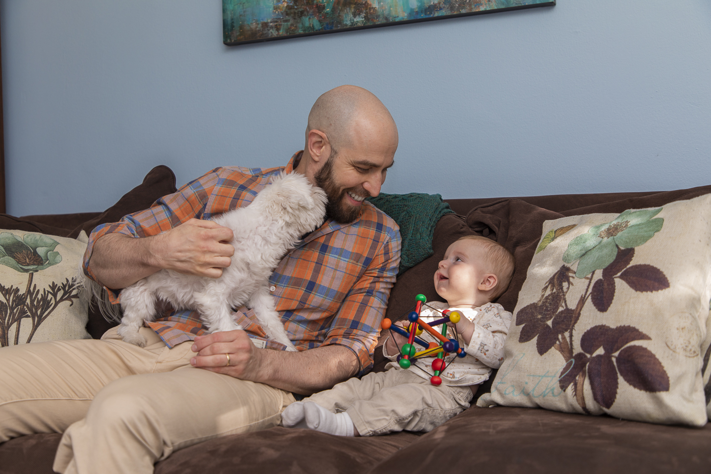 Arthur Migliazza, left, sits on his couch while looking at his newborn baby. Arthur is holding a small dog, and wearing an orange and blue plaid flannel shirt and khaki pants. His newborn is wearing a white onesie and is holding a toy.