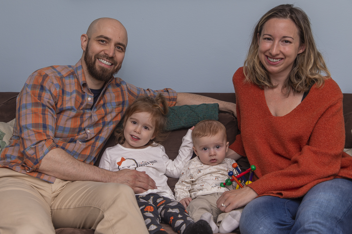 Arthur Migliazza, left, and his wife sit on their couch. Their young daughter and newborn son sit in between them. They are all looking at the camera and smiling. Arthur is wearing an orange and blue plaid flannel shirt and khaki pants. His wife is wearing an orange jumper and blue jeans. Their newborn is wearing a white t-shirt and khaki pants. Their daughter is wearing a white graphic t-shirt and navy graphic pants.