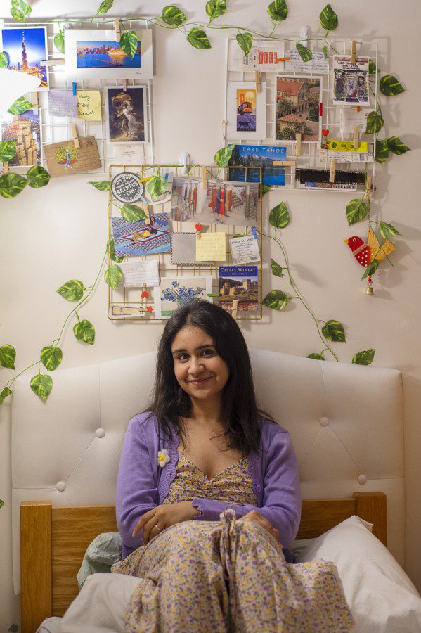 Sara Sharma sits on a dorm room bed, in front of a well-decorated wall with photos and leaf decor hung up. Sara is looking at the camera and wearing a white floral dress and a lilac cardigan.
