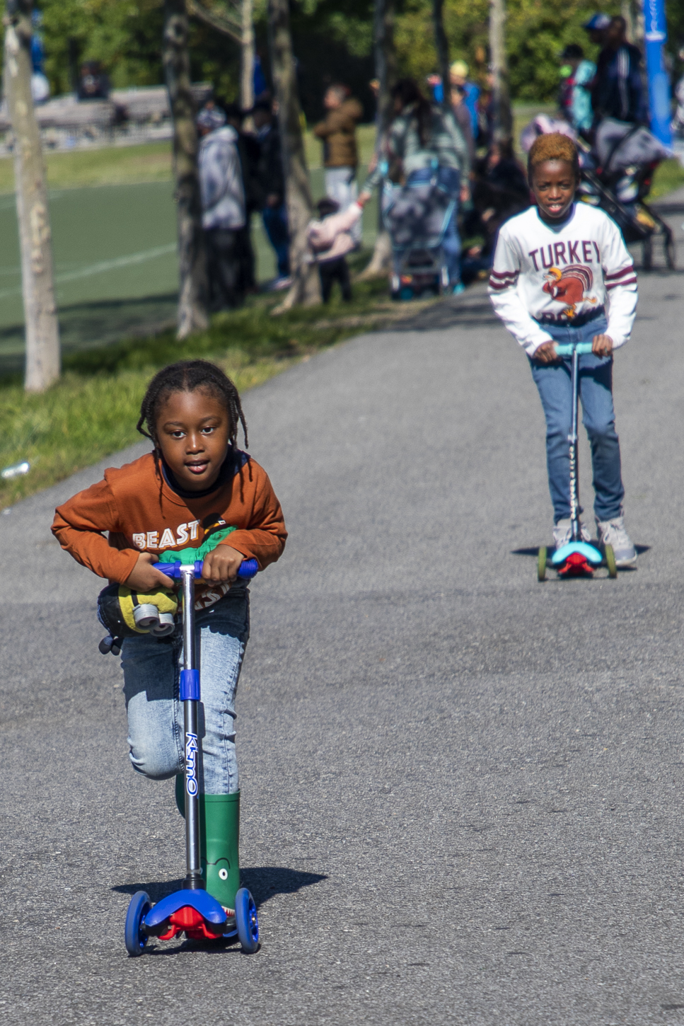 Chauntenay Young’s younger son, to the left, rides a scooter towards the camera as his older brother, to the right, follows along in the background. Her younger son is wearing a graphic orange t-shirt and blue jeans. Her older son is wearing a graphic white t-shirt and blue jeans.