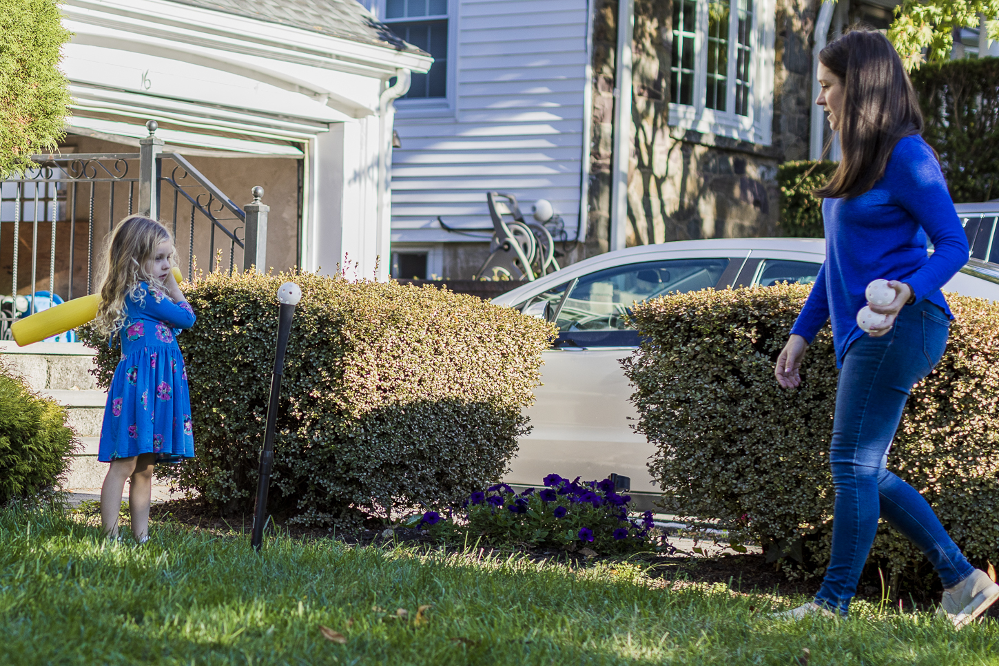 Jessica York, to the right, walks toward her daughter as her daughter hits a softball with a yellow bat. Her daughter is wearing a floral blue dress, and Jessica is wearing a blue shirt and blue jeans. They are standing on the front lawn outside of their home in New Jersey.
