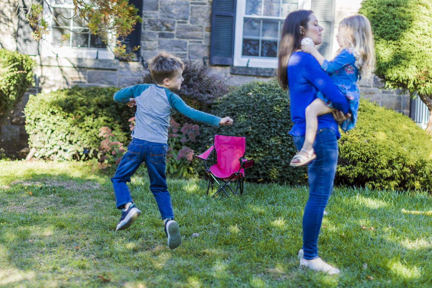 Jessica York stands, holding her daughter as her son (left) spins around, playing in their front yard. Jessica is wearing a blue shirt and blue jeans. Her daughter is wearing a floral blue dress, and her son is wearing a gray and green henley shirt with blue jeans.