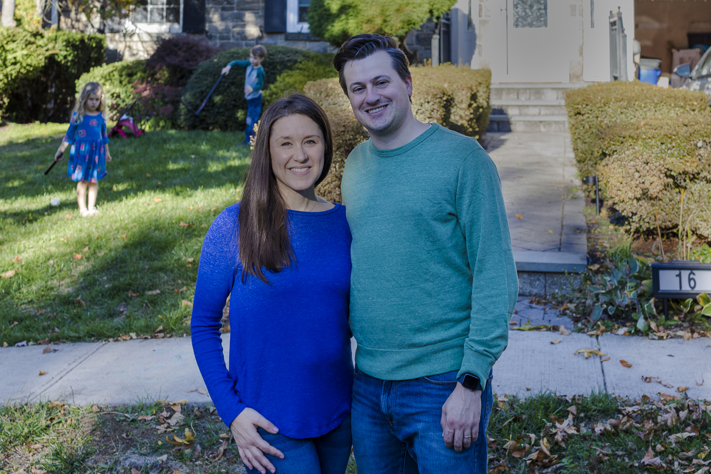 Jessica York poses with her husband in front of the driveway of their home in New Jersey. In the background, their young son and daughter are running around and playing with one another. Jessica is wearing a blue shirt and blue jeans, and her husband is wearing a green sweater and blue jeans. Their daughter is wearing a floral blue dress, and their son is wearing a gray and green henley shirt with blue jeans.