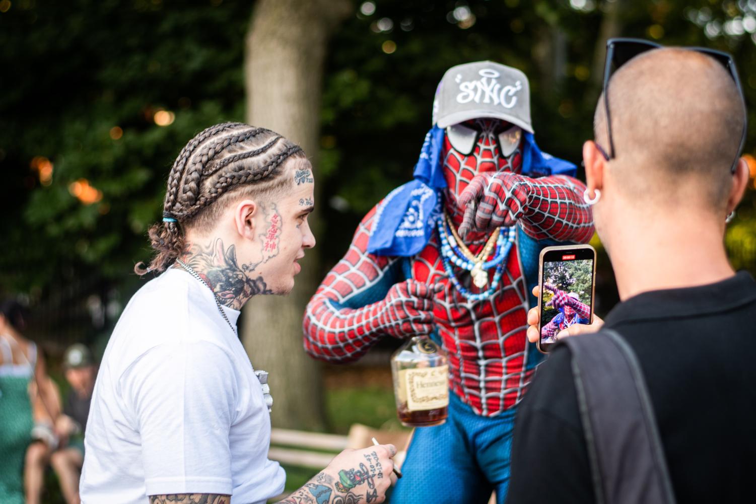 A man in a black shirt records an iPhone video of a man in a red-and-blue Spiderman costume in front of him. To the left, a man with a white shirt and tattoos on his neck, face and arms.