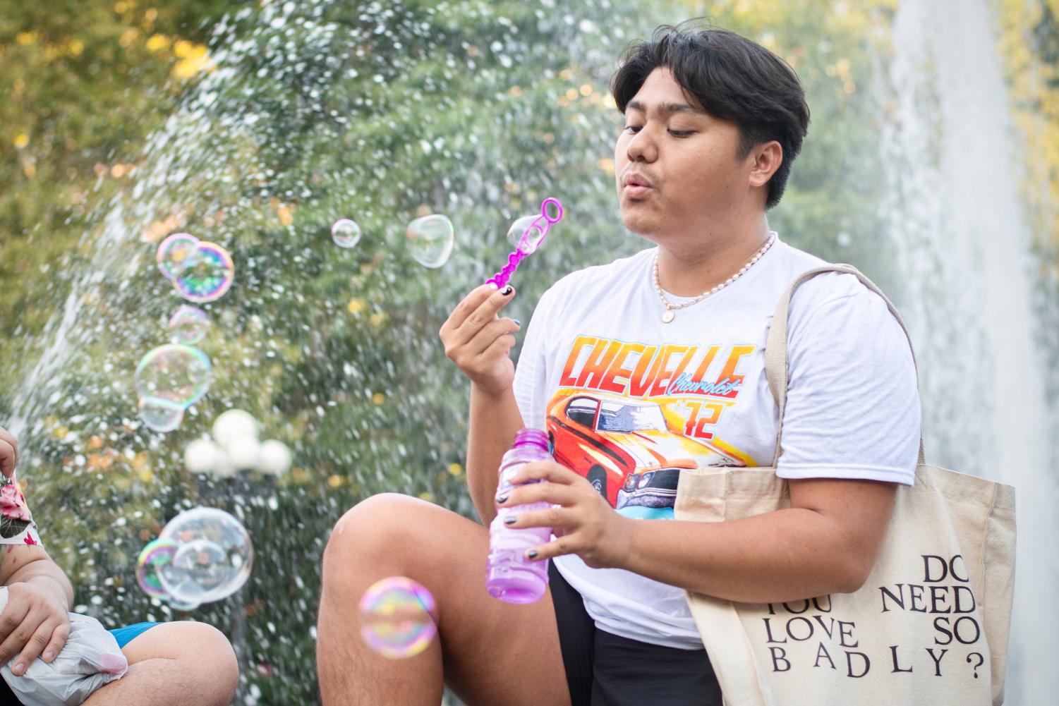 A man wearing a white shirt with an orange car cartoon, black shorts and a tote bag. He is holding a pink bottle of bubbles with one hand and blowing bubbles with his other hand. Behind him is water from a fountain.
