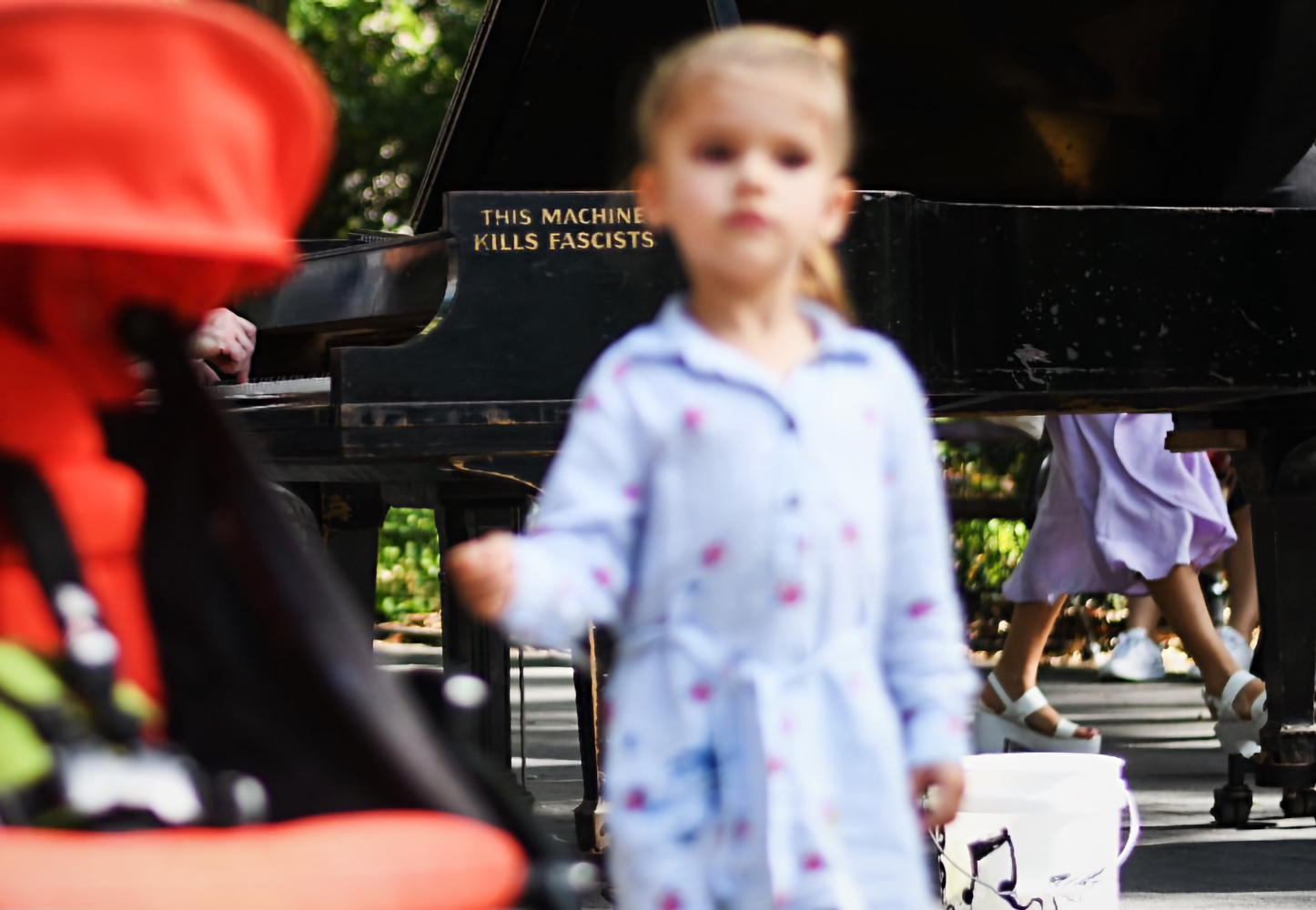 A blonde-haired toddler wearing a blue long-sleeved dress stands in front of a black grand piano that reads in small gold imprint, “This Machine Kills Fascists”.