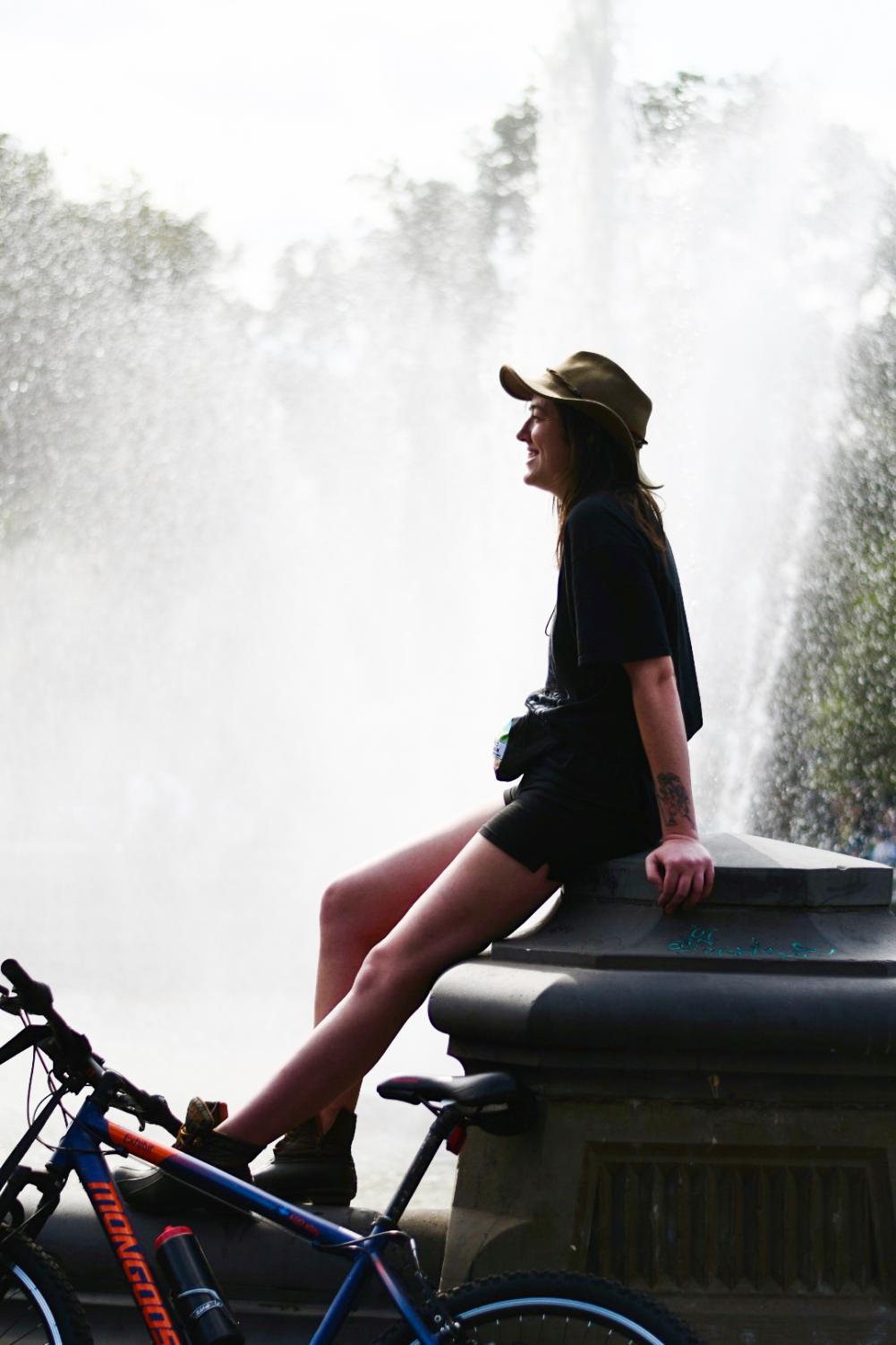 A woman wearing a black shirt, black shorts and a green hat sits on the knee-level concrete structure surrounding the Washington Square Park fountain. To her left is a blue-and-red bicycle.