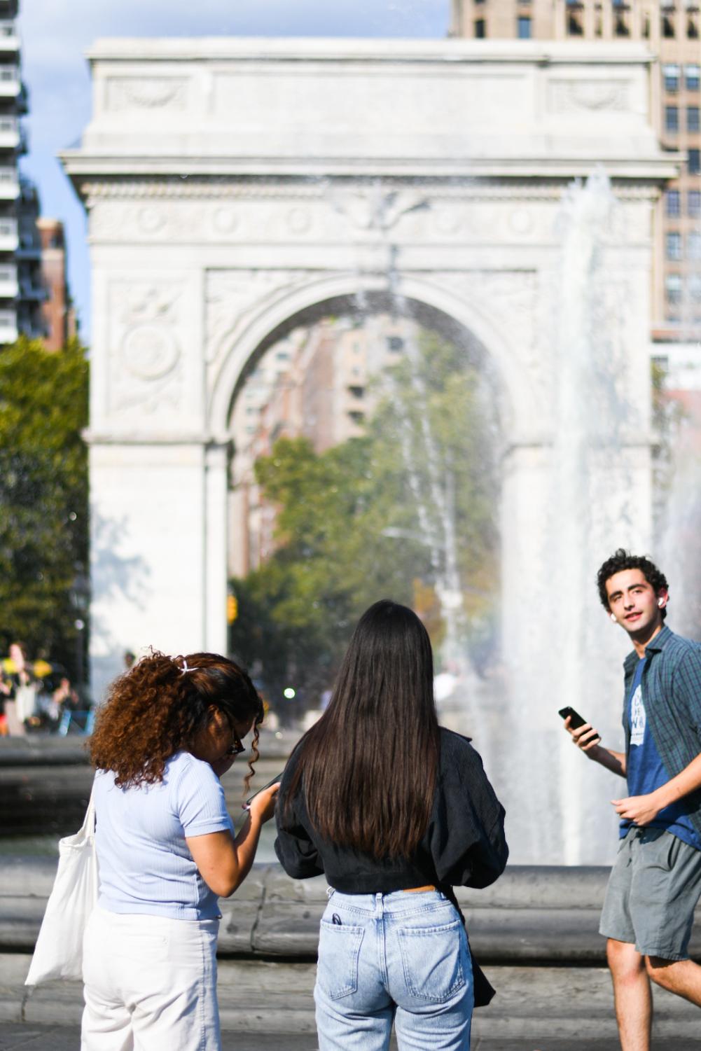 The back of a girl wearing a black shirt and jeans, standing next to another girl wearing a blue shirt and white jeans. Behind them is the marble arch of Washington Square Park, and the park fountain.