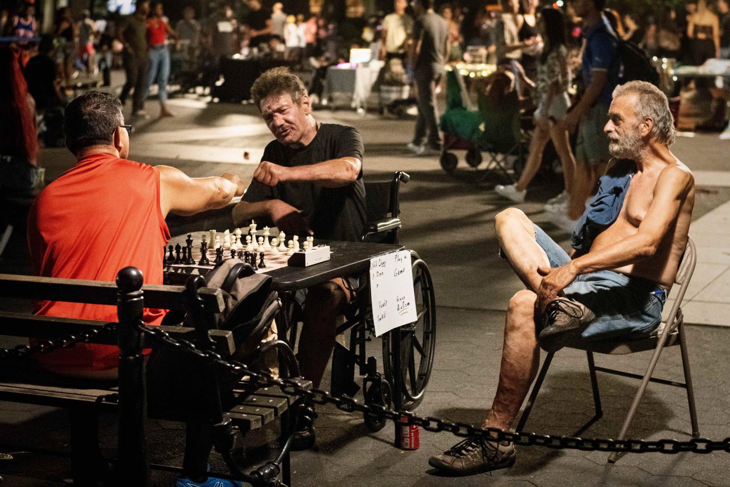 A man wearing a red shirt and a man wearing a black shirt sit facing each other. Between them on a black table is a brown-and-tan wooden chess board. Next to them on a foldable chair sits a man with gray hair, no shirt and blue jean shorts.