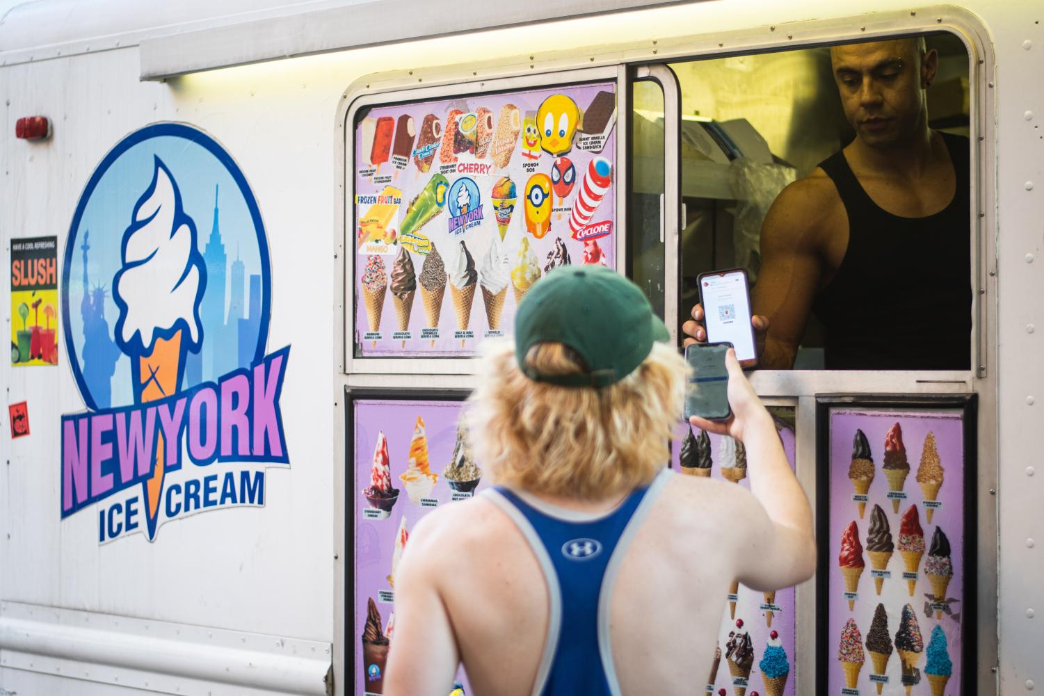 A white ice cream truck with a sign that reads “New York Ice Cream”. Inside the truck a man wearing a black sleeveless shirt holds out his phone to another man wearing a green cap and a blue sleeveless shirt.