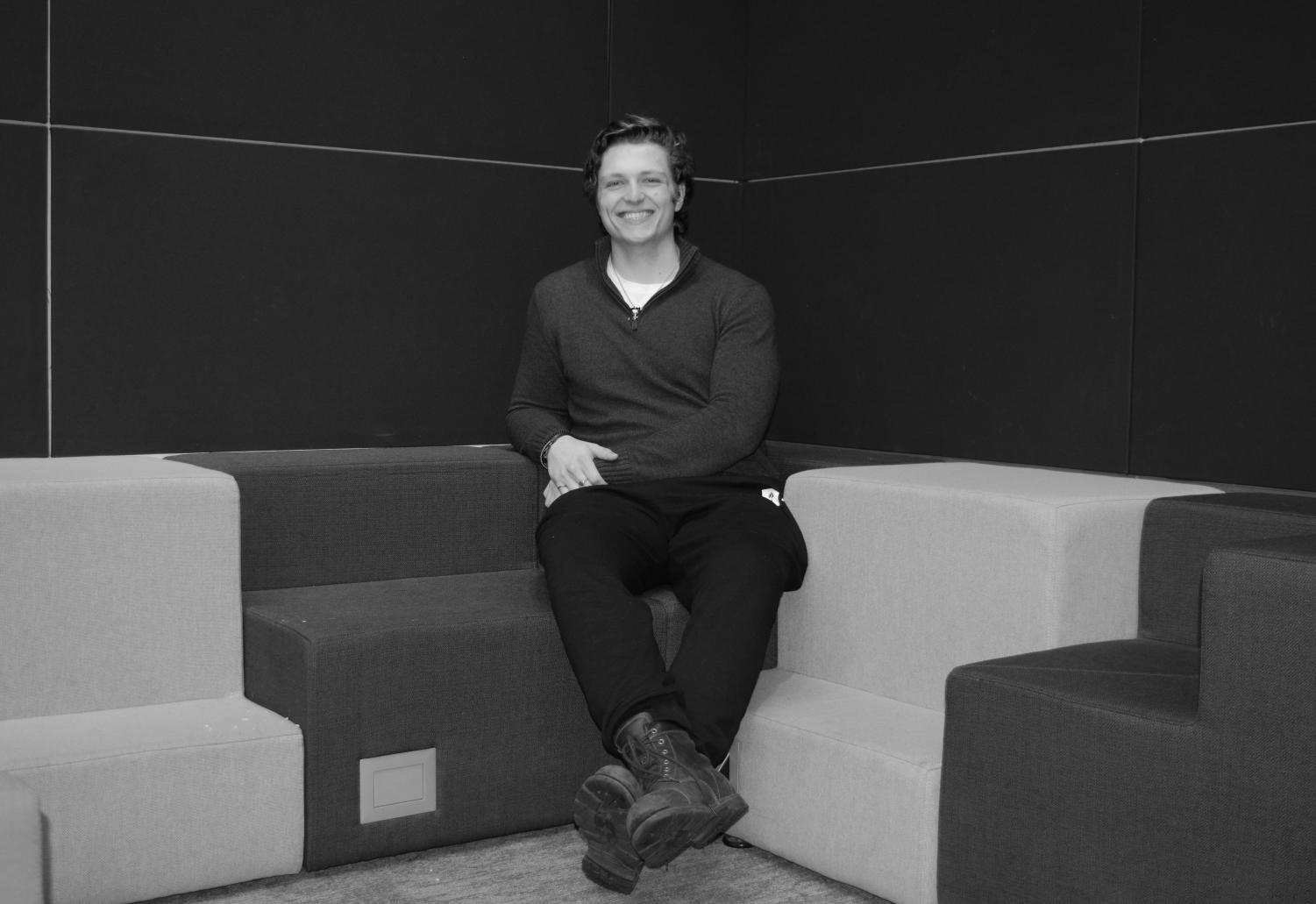 A black-and-white image of a male college student with combed hair, a long-sleeved quarter zip top, dark joggers, and dark boots smiling at the camera while sitting on a couch.