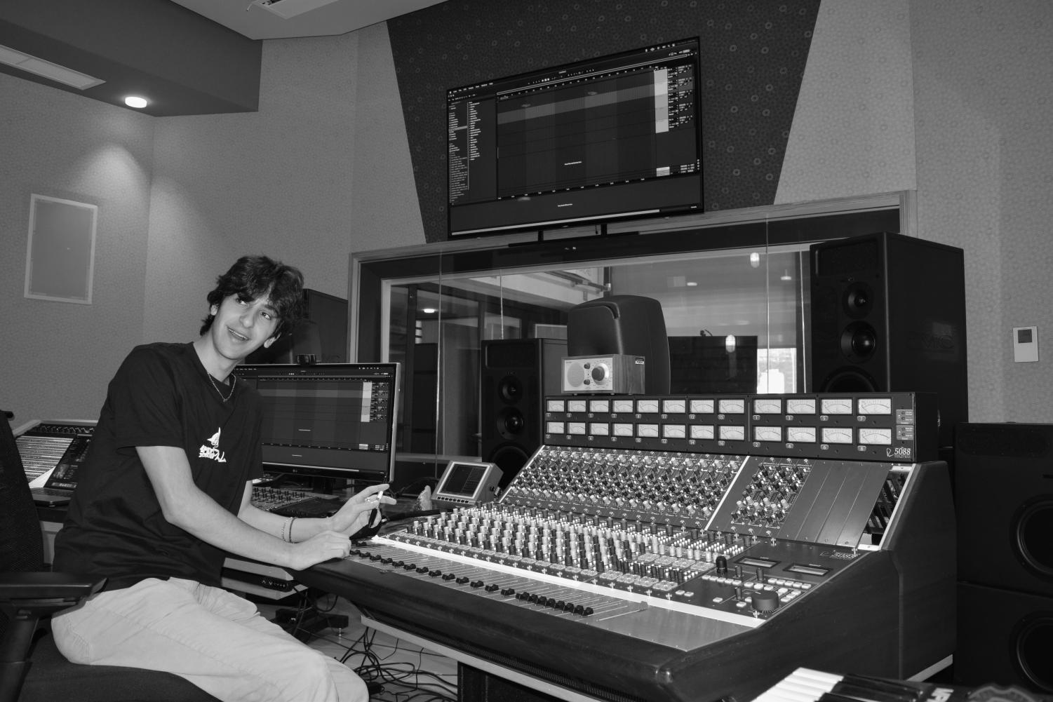 A black-and-white image of a male college student with floppy dark hair wearing a black t-shirt with an unclear logo, light pants, and a small necklace sitting in a chair facing music controls inside a recording studio.