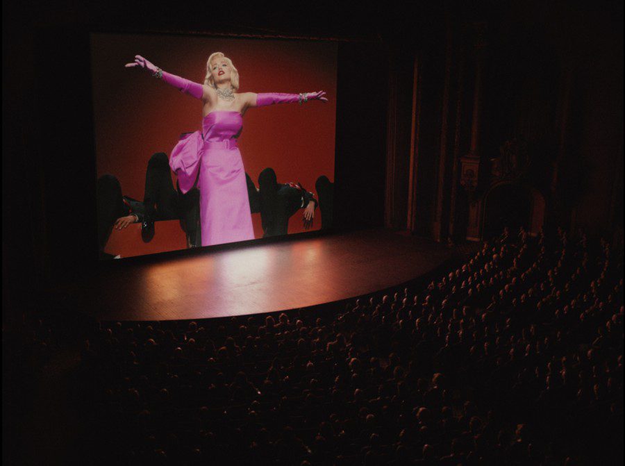 A full crowd of people sit at a movie theater. The screen shows Marilyn Monroe, dressed in a fuschia gown and gloves, gesturing in front of a line of people laying down.