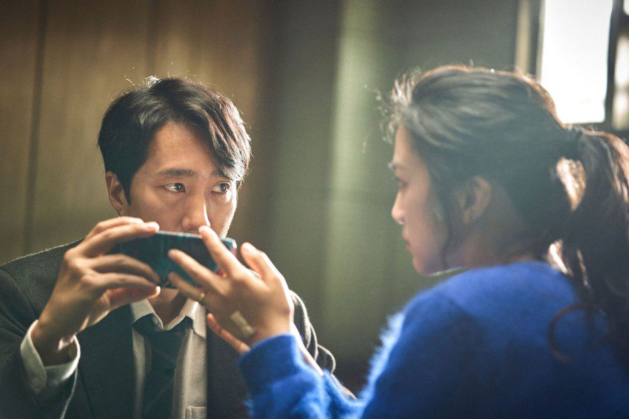 A man wearing a gray suit looking at a woman wearing a blue sweater while they hold a smartphone together.