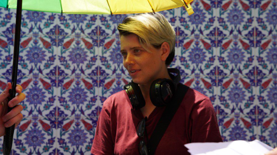 Filmmaker Charlotte Wells wears a red shirt and a pair of black headphones under a colourful umbrella. She stands against a wall with blue patterns.