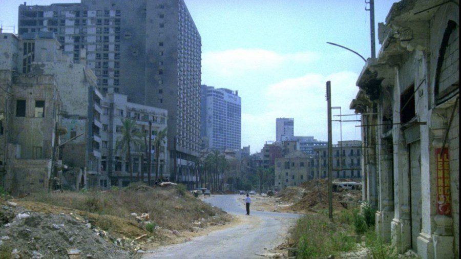 A lone person stands in the middle of a winding road with worn-down buildings and dry vegetation surrounding both sides of the path.