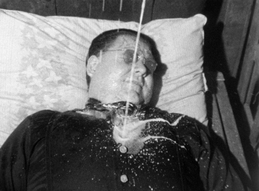 A middle-aged male wearing a pair of sunglasses lies on a bed with white pillow. On the man’s chest is a glass with milk-like white liquid splattering over his clothes.