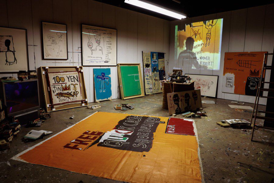An art studio with four framed artworks displayed on the walls and five on the floor. An image with a man wearing a striped shirt writing on a cardboard is projected onto the wall on the right. A ladder, art supplies and several opened books are laying across the studio floor. In the center of the photo is a large canvas with an orange background and “FREE QUICK CASH SHOE BUSINESS” and “RUSH” written on a patch of red paint at the bottom right corner.
