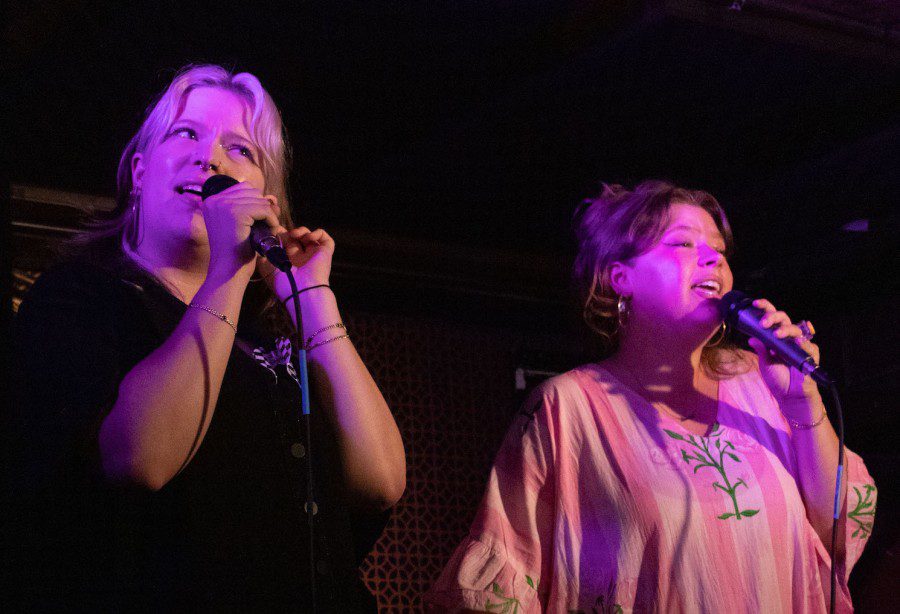 Two twins wear dresses, one black dress and the other white, while standing on stage basking in a purplish spotlight against a dark background. Each holds a microphone and sings.