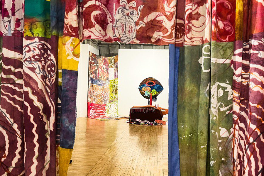 A colorful eyeball-looking art installation sits in the middle of a gallery surrounded by color patterned drapes.