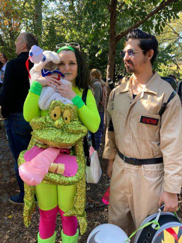 A couple dressed in “Ghostbuster” outfits. The woman on the left dressed in a neon-green monster outfit holds a cat dressed in a white costume. The man on the right wears a beige work jumpsuit.