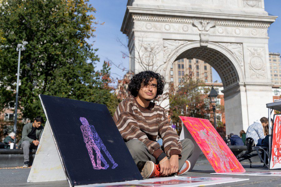 Artist Antonio Garcia, who has black curly hair and is wearing a striped shirt, gray pants and orange shoes, sits on the ground in front of the Washington Square Arch, with a selection of his paintings displayed around him. To his left is a canvas with a dark purple background depicting two human-like figures, the one on the left being bright pink and the one on the right being light purple. To his right is a canvas painted light red with a smeared texture depicting a multicolored cow-like creature.