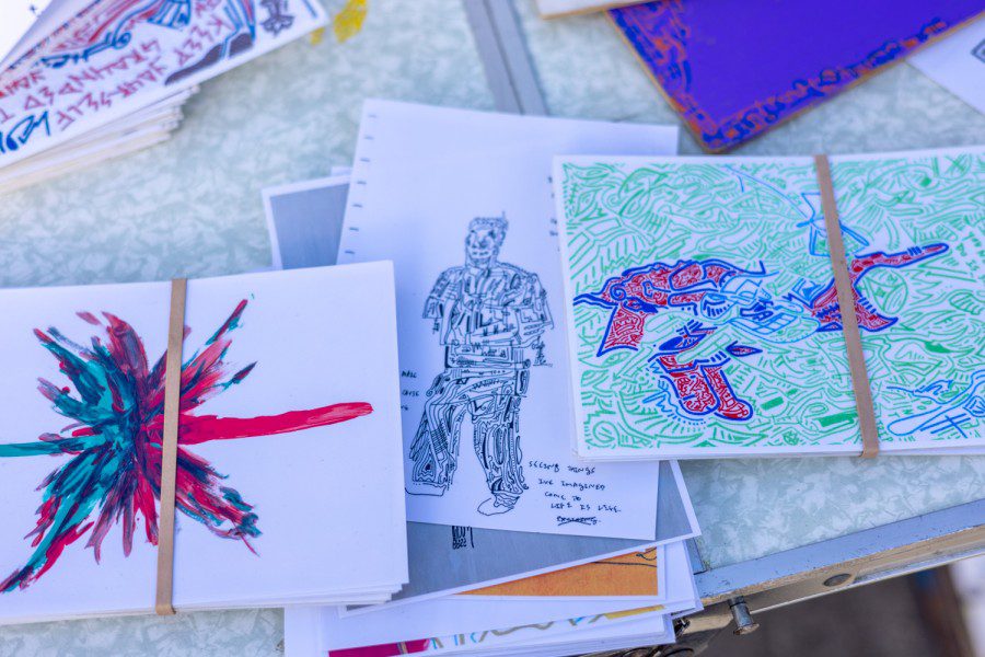 Several stacks of artworks by artist Antonio Garcia laid across a foldable table. On the top of the stack on the left is a white sheet of paper with a turquoise-and-pink butterfly-like creature. On the top of the stack in the center is a white sheet with a row of square holes on the left border, depicting a human-like figure and geometric patterns. On the top of the stack on the right is a sheet of paper covered with a background of turquoise-lined patterns. A red figure sits in the center of the sheet, with the lines making it up bordered in blue.